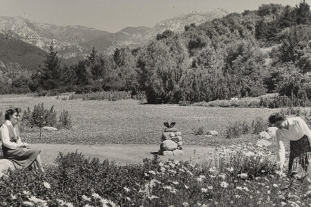 Two women in mid-calf skirts and blouses sit in the meadow. It's a historic moment, captured in black and white, there's a stone tower in the center of a dirt circle. The expansive meadow is just dirt at this time, but the mountains still stand tall in the background and large bushy trees flow down into the garden.