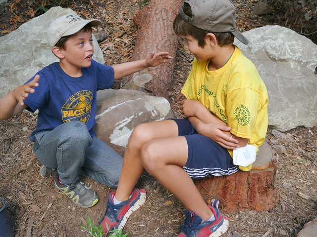 Two smiling kids sitting outside one kid explaining with arms stretched wide