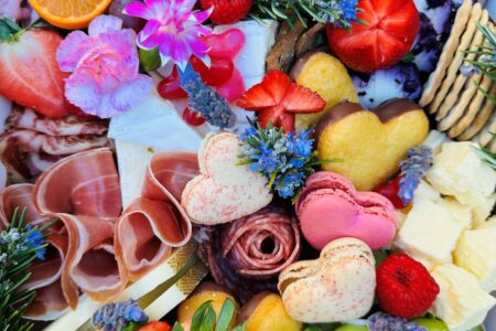 A colorful Charcuterie Mother's Day table full of pink heart shaped macarons, strawberries, flower petals, cheese and crackers, and grapes.