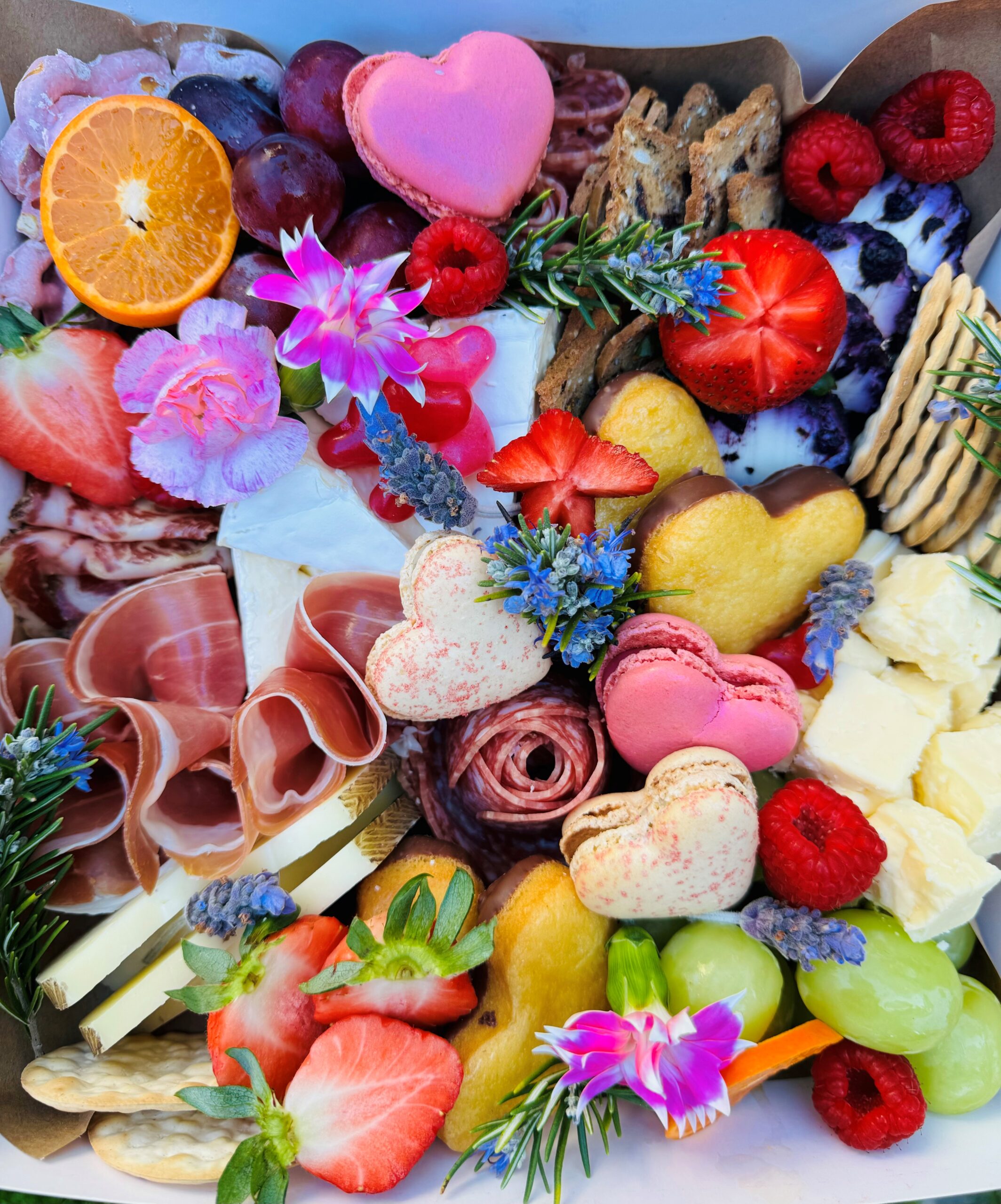 A colorful Charcuterie Mother's Day table full of pink heart shaped macarons, strawberries, flower petals, cheese and crackers, and grapes.