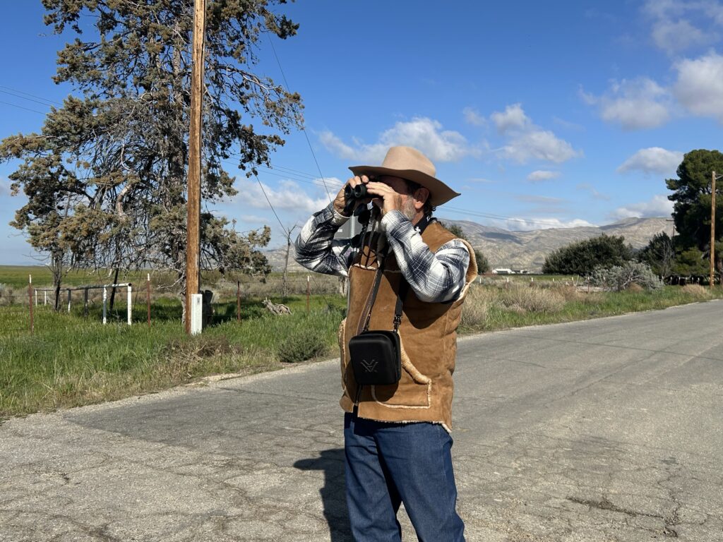 A man standing in a road looking through his binoculars for birds. 