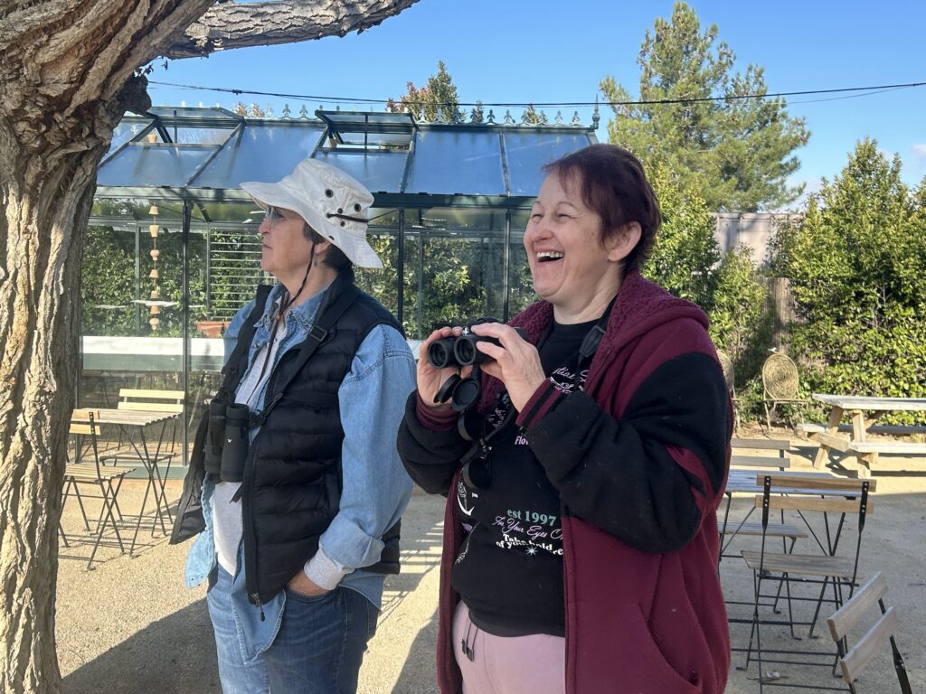 Two women standing under a tree. One woman is smiling after catching sight of a bird in her binoculars. 