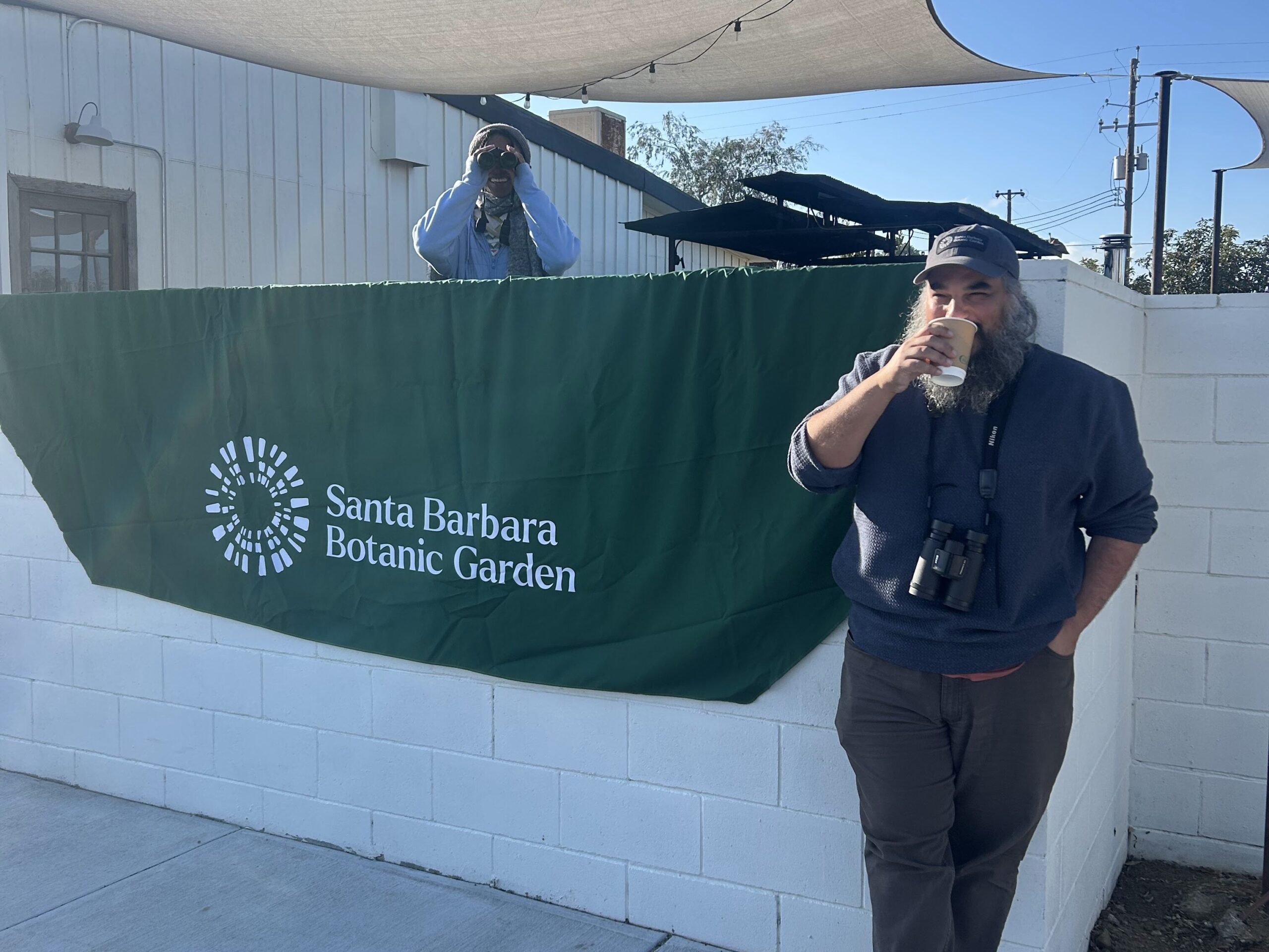 Two Garden staff taking a coffee break. One is the background being funny, hiding behind a wall, looking at the camera man through her binoculars. 